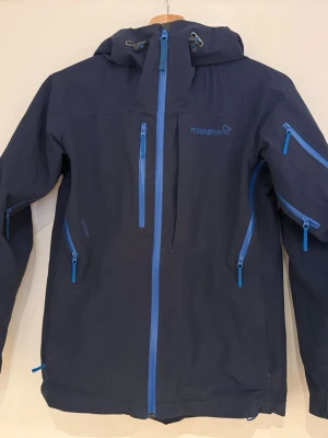 Mörkblå skidjacka från Norrøna strl: 152 - Säljer en mörkblå skidjacka från Norrøna med huva och flera praktiska fickor med blå dragkedjor. Jackan har justerbara ärmslut med kardborre och detaljer som gör den perfekt för utomhusaktiviteter. Materialet är slitstarkt och vattenavvisande syntet. Finns på fler sidor. 