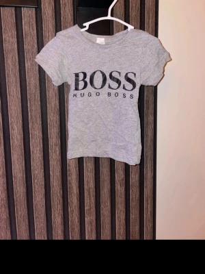 Grå Hugo Boss t-shirt med tryck storlek 68 - Snygg grå t-shirt från Hugo Boss med stort svart BOSS-tryck på bröstet. Klassisk passform och rund hals. Tillverkad i mjuk bomull som känns skön mot huden. Perfekt för dig som gillar stilrena och enkla plagg med logga. Storlek 68