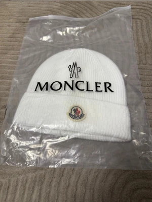 Vit Moncler mössa i ull - Säljer en stilren vit Moncler mössa i ribbstickad ull med klassisk Moncler-logga framtill. Mössan har vikt kant och är perfekt för dig som vill ha en clean och exklusiv look under kalla dagar.