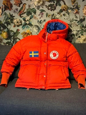 Acne Studios x Fjällräven Orange Expedition dun  - Säljer en fet röd dunjacka från Acne Studios x Fjällräven med stor huva, blått innerfoder och coola detaljer som svenska flaggan och logopatch. Jackan har stora fickor, knappar med branding, dragkedja och snörning vid huvan. Perfekt för kalla dagar och sticker ut med sin färg och design.