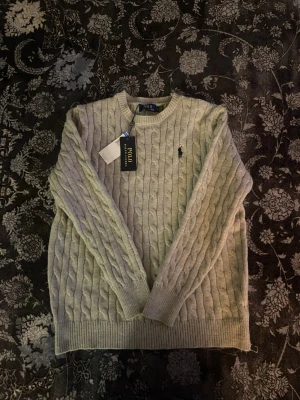 Grå stickad tröja från Polo Ralph Lauren - Snygg grå kabelstickad tröja från Polo Ralph Lauren med klassisk rund hals och broderad logga på bröstet. Tröjan har långa ärmar och ribbade muddar vid ärmslut och nederkant. Perfekt för dig som gillar stilrena och tidlösa plagg.