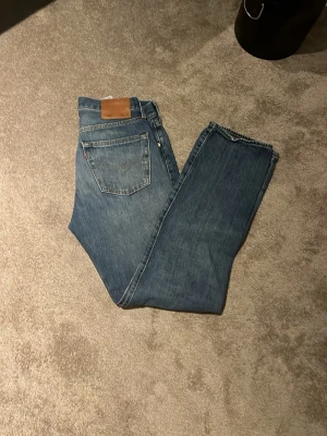 Levi's 501 jeans blå W30 L32 - Klassiska Levi's 501 jeans i blå tvätt med raka ben och knappgylf. Jeansen har fem fickor, brunt läderpatch bak och subtila slitningar framtill. Tillverkade i slitstarkt bomullsjeansmaterial, perfekt för en chill och tidlös look.