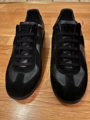 Maison Margiela  - Maison Margiela sneakers i helsvart med snygga paneler i mocka och skinn. Klassisk rund tå och platt sula, med svarta snören och diskret logga på tungan. Perfekt för dig som gillar minimalistisk och stilren design.