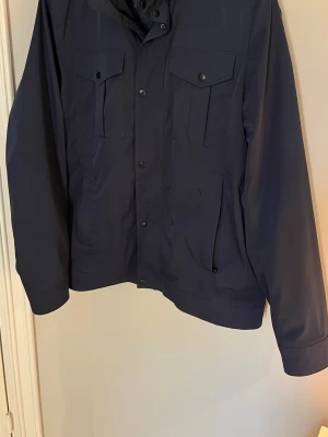 Marinblå field jacka från J.Lindeberg - Snygg marinblå vindjacka från J.Lindeberg , den populära modellen: baylie poly feild jacket. med hög krage, tryckknappar och dragkedja. Jackan har två bröstfickor med lock och två sidofickor. Perfekt för dig som gillar stilren och funktionell design.