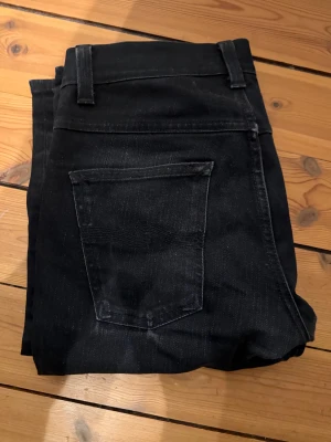 Nudie Jeans - Säljer ett par svarta skinny jeans från Nudie Jeans i storlek 29/32. Jeansen har klassisk femficksdesign, smal passform och är tillverkade i stretchig bomull. Snygga detaljer med svarta sömmar och Nudie Jeans-logga på bakfickan.