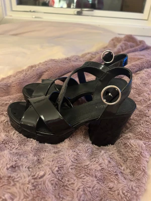 Svarta sandaler med klack från Divided - Säljer ett par svarta sandaler med breda, glansiga remmar och rundad öppen tå. Skorna har hög klack och plattform, samt silverfärgade spännen vid ankeln. Materialet är syntet med lackad finish som ger en cool vibe.
