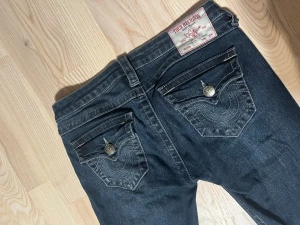 True Religion Lågmidja Bootcut Jeans - Säljer ett par mörkblå jeans från True Religion med klassiska sömmar och snygga fickdetaljer med knappar bak. Modellen har bootcut och låg midja, tillverkade i jeansmaterial med en cool tvätt. Strl 24, passar mig som är 164cm lång med marginal. 🥰