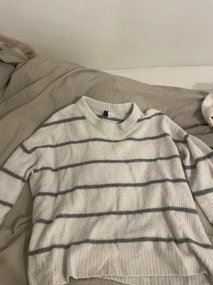 Randig stickad tröja från H&M Divided - Mysig stickad tröja från H&M Divided i storlek M. Tröjan är vit med horisontella grå ränder och har en rund halsringning samt långa ärmar. Perfekt för kyliga dagar och enkel att matcha med jeans eller kjol.