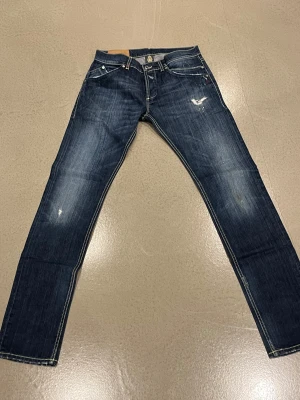 Dondup jeans med slitningar - Säljer ett par mörkblå dondup  med snygga slitningar och detaljerad brodering vid fickan. Jeansen har klassisk femficksdesign, kontrastsömmar och är tillverkade i slitstarkt bomullsjeansmaterial. Passformen är slim och midjan är normal.