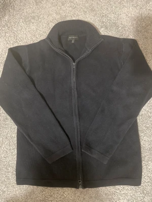 Svart stickad kofta från Steel - Svart stickad kofta från Steel i storlek S. Koftan har hel dragkedja framtill, ribbad krage och långa ärmar. Perfekt för lager-på-lager under kyliga dagar. Enkel och stilren design som funkar till många olika outfits.