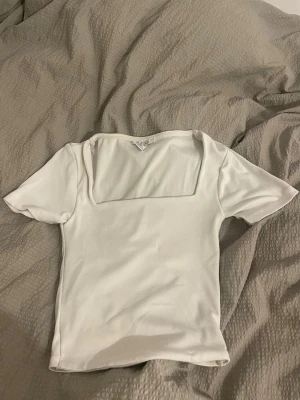 Vit topp med fyrkantig halsringning - Säljer en vit topp från New yorker med korta ärmar och en snygg fyrkantig halsringning. Toppens passform är normal och materialet känns mjukt mot huden. Perfekt att styla med jeans eller kjol för en clean look.