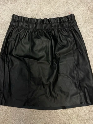 Svart skinnkjol från Gina Tricot - Säljer en svart kort skinnkjol från Gina Tricot i storlek M. Kjolen har en elastisk midja som ger en bekväm passform och en stilren look. Perfekt för dig som gillar att sticka ut med en edgy vibe. Materialet är syntetiskt skinn som känns mjukt och följsamt.