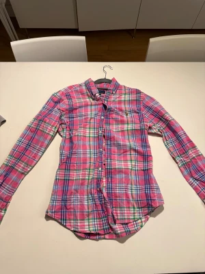 Rutig rosa skjorta från Ralph Lauren - Säljer en slim fit skjorta från Ralph Lauren i storlek M. Skjortan har ett färgglatt rutigt mönster i rosa, blått, grönt och vitt, klassisk krage och långa ärmar. Tillverkad i mjuk bomull och har knappar framtill. Perfekt för dig som gillar färg och stil.