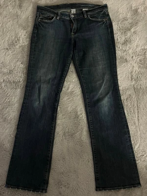 Lucky Brand bootcut jeans mörkblå - Säljer ett par mörkblå bootcut jeans från Lucky Brand med klassisk femficksdesign och snygga slitningar. Jeansen har gul kontrastsöm, läderpatch bak och dragkedja framtill. Modellen heter Sundown Jean och är tillverkad i bomull.    Mått: midja-41cm(tvärs över) grenhöjd-22. Innerbenslängd-80cm.  Benöppning- 23cm. Totallängd- 103cm