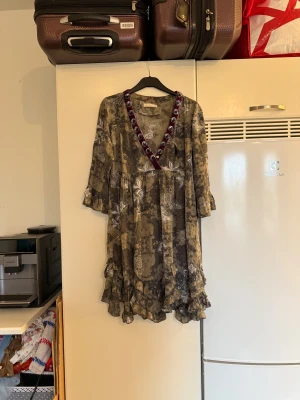 Odd Molly mönstrad kortklänning - Supersöt kortklänning från Odd Molly i grönt och grått med vitt blommigt mönster. V-ringad med lila sammetskant och silverfärgade paljetter. Volangdetaljer nedtill och på ärmarna, trekvartsärm och loose passform. Perfekt för dig som gillar boho-vibe! Från märket Odd Molly. Storlek 2 som enligt deras storleksguide motsvarar en M. Mycket fint skick, jag hittar inga synliga anmärkningar eller defekter.  