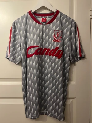 Liverpool tröja - Retro Liverpool fotbollströja i grått med röd logga och Candy-tryck på bröstet. Tröjan har ett unikt rutigt mönster och röda detaljer på krage och axlar. Perfekt för dig som gillar klassisk fotbollsstil och vill sticka ut på läktaren.