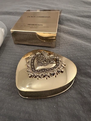 Dolce & Gabbana Ever Icon Eye Palette - Lyxig ögonskuggspalett i hjärtformad guldförpackning med dekorativt emblem. Paletten 'Amber Venus' innehåller sex nyanser i varma bruna och gyllene toner, både matta och skimrande. Spegel ingår i locket. Perfekt för att skapa snygga ögonlooks både för vardag och fest. 