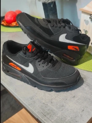 Nike Air Max 90 svart och vit - Nike Air Max 90 sneakers i svart med vita swoosh-detaljer och orange Air Max-logga på sidan. Skorna har snörning, mesh och skinnpaneler samt synlig luftdämpning i sulan. Perfekt för dig som gillar klassisk streetstil och vill ha bekväma sneakers med snygg design.
