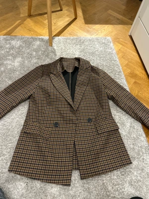 Brun rutig kavaj med knappar - Snygg kavaj i brunt och svart rutigt mönster med klassisk krage och två framfickor. Dubbelknäppt design med svarta knappar och helfodrad insida. Perfekt för dig som gillar en stilren och trendig look. S/M