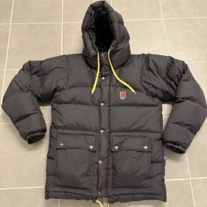 Svart pufferjacka från Fjällräven XS - Svart pufferjacka från Fjällräven med huva och gula snören. Jackan har två stora fickor med knappar framtill, dragkedja och tryckknappar. Klassisk Fjällräven-logga på bröstet och quiltad design. Perfekt för kalla dagar.
