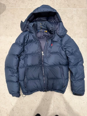 Mörkblå dunjacka från Ralph Lauren - Mörkblå dunjacka från Polo Ralph Lauren, köpt på NK i Stockholm för ca 5300 kr. Klassisk och stilren modell med huva, bra passform och riktigt varm – perfekt för höst och vinter. Storlek S.  Skick: suveränt skick, väldigt sparsamt använd. Inga hål, fläckar eller defekter.  Säljer då jag har köpt en annan jacka och denna inte kommer till användning, därför lågt pris. Kvitto finns ej, men äkthet garanteras.  Mitt pris: 1700 kr Hör gärna av dig vid frågor eller om du vill ha fler bilder.