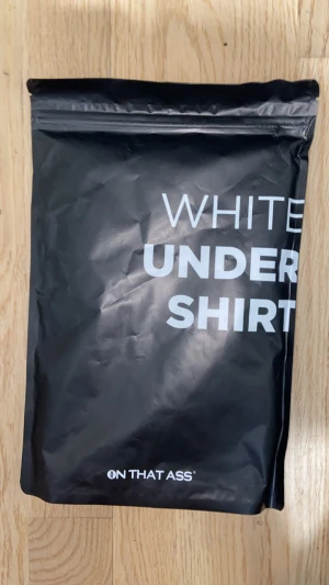 Vit undershirt från ON THAT ASS - Fräsch vit undershirt från ON THAT ASS, perfekt att ha under andra plagg. T-shirten har klassisk rund hals och korta ärmar, med en clean och enkel design. Materialet är mjukt och skönt, troligtvis bomull eller polyester för maximal komfort.