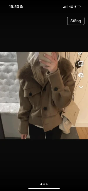 Beige jacka med päls från Meotine - Säljer en beige jacka från Meotine med fluffig päls runt kragen. Jackan har stora svarta knappar, två bröstfickor med lock och en loose passform. Materialet är ullblandning och pälsen ger en lyxig vibe. Perfekt för dig som vill ha en snygg och trendig look.