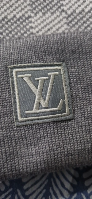 Grå rutig mössa med L v-logga - Grå mössa från L v med diskret rutmönster och uppvikt kant. Framtill sitter en vit patch med svart L v-logga. Mössan är stickad och har en exklusiv känsla, perfekt för dig som vill ha en clean och lyxig look.