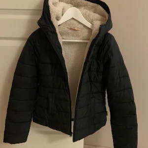 Svart pufferjacka med teddyfoder - Svart pufferjacka från Hollister med mysigt teddyfoder i beige. Jackan har huva, dragkedja framtill och två sidofickor. Quiltad design och långärmad modell som ger en chill och varm vibe. Perfekt för kalla dagar när du vill vara både bekväm och snygg. Sytt själv vid axeln i innerfodret, det syns lite (ej utifrån) men man kan lätt sy bättre själv om man vill att det inte ska synas.