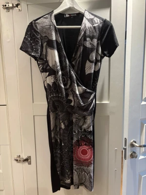 Mönstrad omlottklänning från Desigual - Säljer en cool omlottklänning från Desigual i svart, grått och vitt med blommigt mönster och en röd detalj. Klänningen har korta ärmar och v-ringad hals. Materialet känns mjukt och stretchigt, perfekt för dig som gillar unika prints och snygg passform.