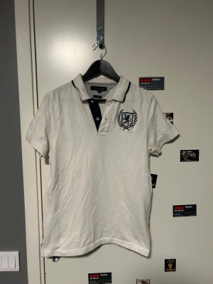 Vit piké från Tommy Hilfiger - Vit pikétröja från Tommy Hilfiger med mörkblå detaljer på krage och knappslå. Broderat emblem med lejon och lagerkrans på bröstet samt liten Hilfiger-logga på ärmen. Klassisk pikékrage och korta ärmar, tillverkad i bomull.