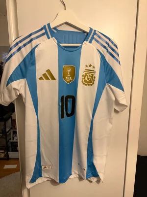 Argentina Messi matchtröja Adidas - Säljer en officiell Argentina landslagströja från Adidas med Messi 10 på ryggen. Tröjan har klassiska blå och vita ränder, guldiga detaljer och FIFA-märke på bröstet. Perfekt för fotbollsfans som vill sticka ut på läktaren eller hemma.
