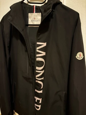 Svart vindjacka från Moncler - Svart vindjacka från Moncler med stor logotyp tryckt längs framsidan och klassisk Moncler-patch på ärmen. Jackan har huva, dragkedja och snörning nertill. Snygg och stilren design med diskreta detaljer och praktiska fickor. 