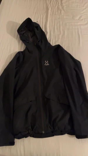 Svart vindjacka från Haglöfs - Svart vindjacka från Haglöfs med huva och hel dragkedja framtill. Jackan har två stora fickor med lock och justerbar nederkant. Perfekt för blåsiga dagar och outdoor-aktiviteter. Materialet är syntet och jackan har en stilren look.