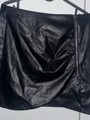 Svart skinnkjol från Threadbare - Säljer en svart kort kjol i skinnimitation från Threadbare. Kjolen har en snygg omlott-design med dragkedja på sidan som ger en cool touch. Perfekt för dig som gillar stilrena och edgy plagg. Passformen är normal och materialet känns mjukt och följsamt.