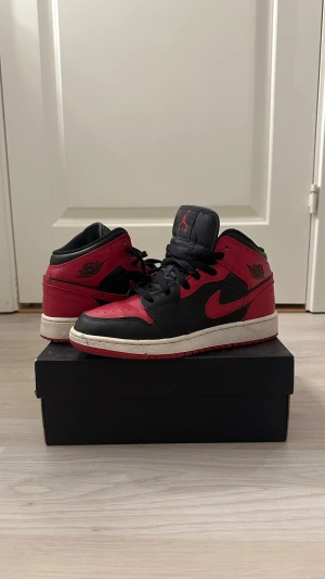 Nike Air Jordan 1 Mid (gs) Svart/Röd - Nike Air Jordan 1 Mid (gs) sneakers i svart och rött med klassisk Wings-logga på sidan. Köpte för några år sedan men nästan inte använda. Är i väldigt bra skick, säljer på grund av att de inte kommer till användning. Original sko låda följer även med. 