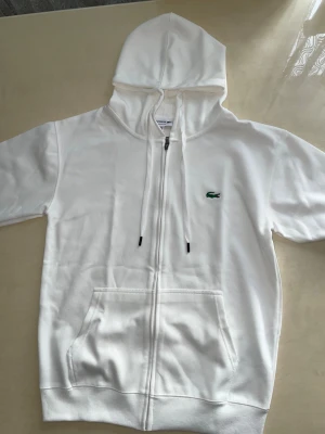 Lacoste-hoodie - Fräsch vit Lacoste-hoodie med dragkedja. Tillverkad i mjukt och kvalitativt material med den ikoniska gröna krokodil-loggan på bröstet. Modellen har justerbar huva och praktisk känguruficka,