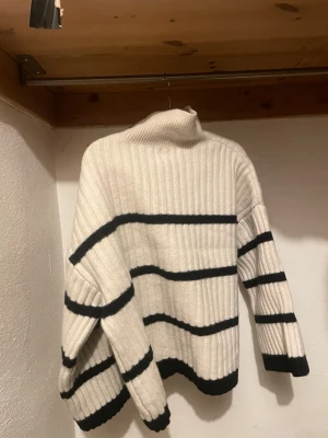 Vit polotröja med svarta ränder - Säljer en ribbad polotröja i vit färg med svarta horisontella ränder. Tröjan har lång ärm och hög krage som ger en chill vibe. Materialet är stickat och känns mjukt och varmt, perfekt för kyliga dagar. Passformen är loose och ger en avslappnad look.