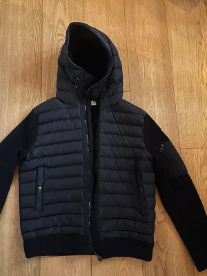 Svart Moncler pufferjacka med huva - Svart pufferjacka från Moncler med huva och ribbstickade ärmar. Jackan har dragkedja framtill, sidofickor med knapp och en extra ficka med dragkedja på ärmen. Materialmix av dun och stickad polyester ger en schysst look och värme.