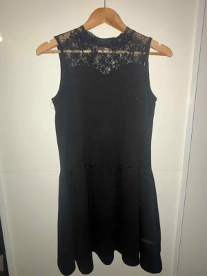 Svart kort klänning med spets - Snygg svart klänning med ärmlös design och spetsdetaljer upptill vid halsen. Kjolen är lätt utställd och klänningen har en feminin siluett som passar perfekt för dig som gillar stilrena plagg med en twist. Perfekt för dig som vill ha något enkelt men ändå elegant.