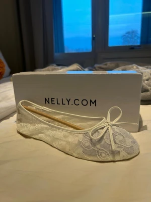 Vita spetsballerinaskor från Nelly - Säljer ett par vita ballerinaskor från Nelly med rund tå och dekorativ rosett framtill. Skorna är täckta med blommig spets och har en platt sula. Jag har aldrig använt dem, helt nya. Säljer pga att dem ej passa