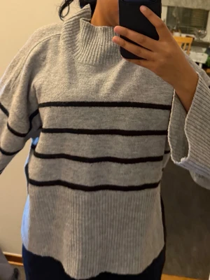 Grå randig stickad tröja med slits - Mysig grå stickad tröja med svarta horisontella ränder och hög krage. Tröjan har långa ärmar och en snygg slits i sidan som ger en cool detalj. Perfekt för lager-på-lager under hösten och vintern.