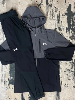 Ingen sko på bilden - Bilden visar ett träningsset från Under Armour med en grå och svart hoodie samt svarta träningsbyxor, men inga skor finns med på bilden.