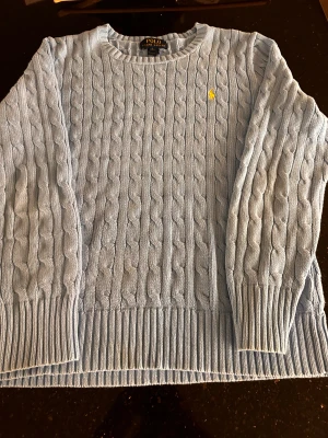 Ljusblå stickad tröja från Polo Ralph Lauren - Säljer en ljusblå stickad tröja från Polo Ralph Lauren i storlek S. Tröjan har klassiskt kabelstickat mönster, rund hals och långa ärmar. På bröstet finns den ikoniska gula Polo-loggan broderad. Perfekt för dig som gillar stilrena och tidlösa plagg.