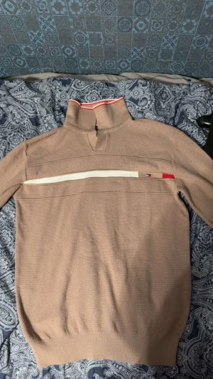 Beige stickad långärmad från Tommy Hilfiger - Säljer en beige stickad långärmad från Tommy Hilfiger med hög krage och half zip. Västen har en vit horisontell rand över bröstet och en liten logotyp i rött, vitt och blått. Kragen har en snygg röd och vit detalj upptill. Den är slim fit så den passr både S och M