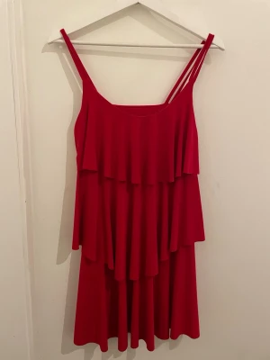 Röd volangklänning med smala axelband - Säljer en röd kortklänning med flera lager volanger och smala axelband. Klänningen har en rundad halsringning och är ärmlös, vilket ger en luftig och flowy känsla. Perfekt för dig som gillar färgstarka och lekfulla plagg.