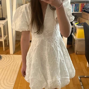 Studentklänning: Vit kort klänning med puffärmar - Superfin vit kort klänning/studentklänning med stora puffärmar och v-ringning. Klänningen har ett broderat blommigt mönster över hela tyget och har en puffig kjol. 🤍