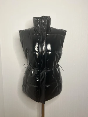 Svart streetstyle dunväst från H&M - Svart dunväst från H&M i storlek S med glansig finish och hög krage. Västen har dragkedja och tryckknappar framtill, två fickor och snörning i midjan för att justera passformen. Perfekt för att ge din outfit en cool edge.