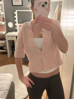 Ljusrosa kofta från Gina Tricot - Supersöt ljusrosa kofta från Gina Tricot med korta ärmar och rund hals. Koftan är ribbstickad och har knappar framtill. Perfekt att slänga över ett linne för en chill look. Mjuk och skön, passar till både jeans och kjol.