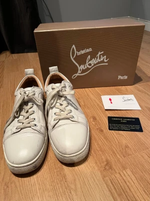 Vita sneakers från Christian Louboutin - Säljer ett par vita sneakers från Christian Louboutin med klassisk röd sula och snygga snörningsdetaljer. Skorna är tillverkade i skinn och har rund tå samt platt sula. Kommer med originalkartong och certifikat. Perfekt för dig som gillar exklusiva märken och stilren design.